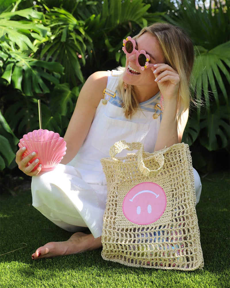 girl next to woven tote bag with eternal optimist smiley holding shell shaped sipper wearing floral sunglasses