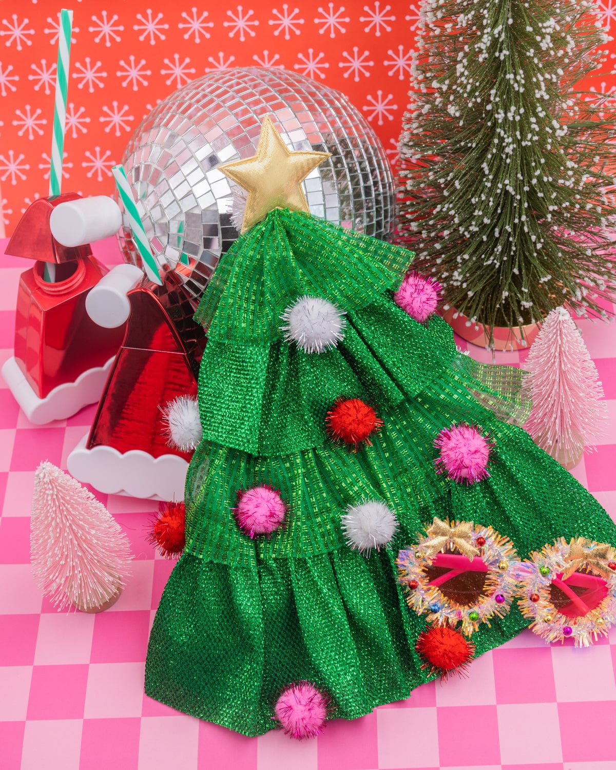 Christmas tree hat placed with assortment of holiday products and decor shot against pink checkered background.