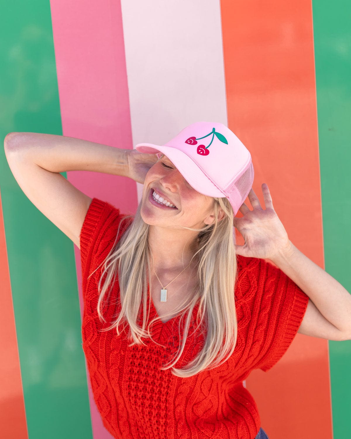 blonde girl smiling and laughing with very cherry trucker hat in front of colorful striped wall
