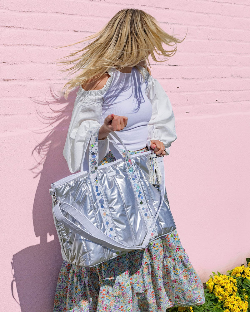 Blonde girl against pink wall with a silver duffle bag with iridescent confetti straps and a white detachable shoulder strap.