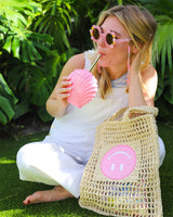 Girl drinking out of pink shell shaped cup with gold straw and woven rattan bag with pink smiley face