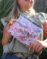 Packed Party Everyday Pouch lifestyle closeup picture. Girl wearing sage green top, holding confetti makeup and toiletry pouch. Navy zipper, gold, red, orange, pink and holographic confetti mix.  