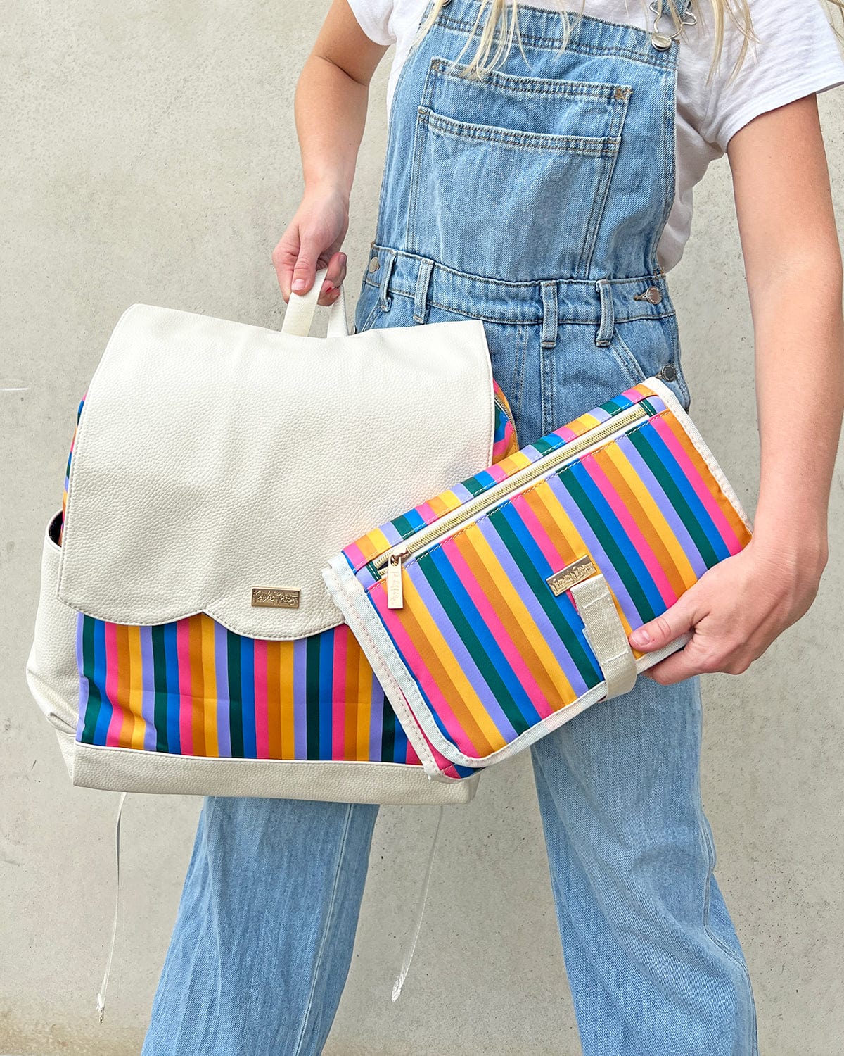 Girl in denim overalls holding cream colored diaper bag backpack with rainbow stripes and rainbow baby changing pad