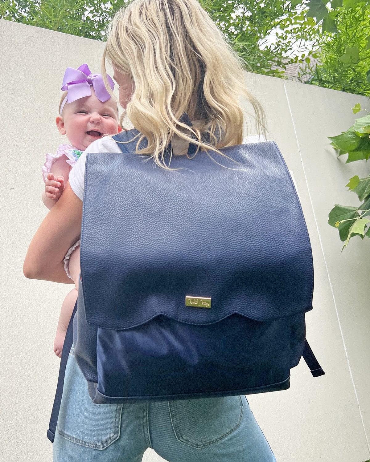 Blonde woman wearing navy diaper bag backpack and holding baby girl with big purple bow