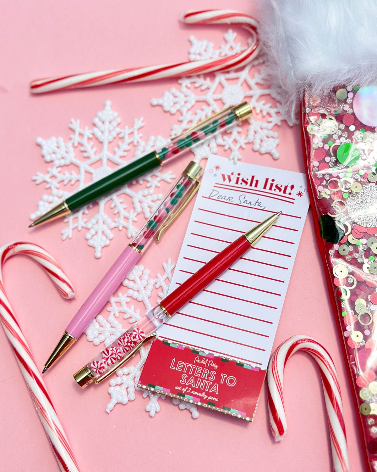 3 festive holiday confetti filled pens surrounded by holiday decor shot against pink background. 
