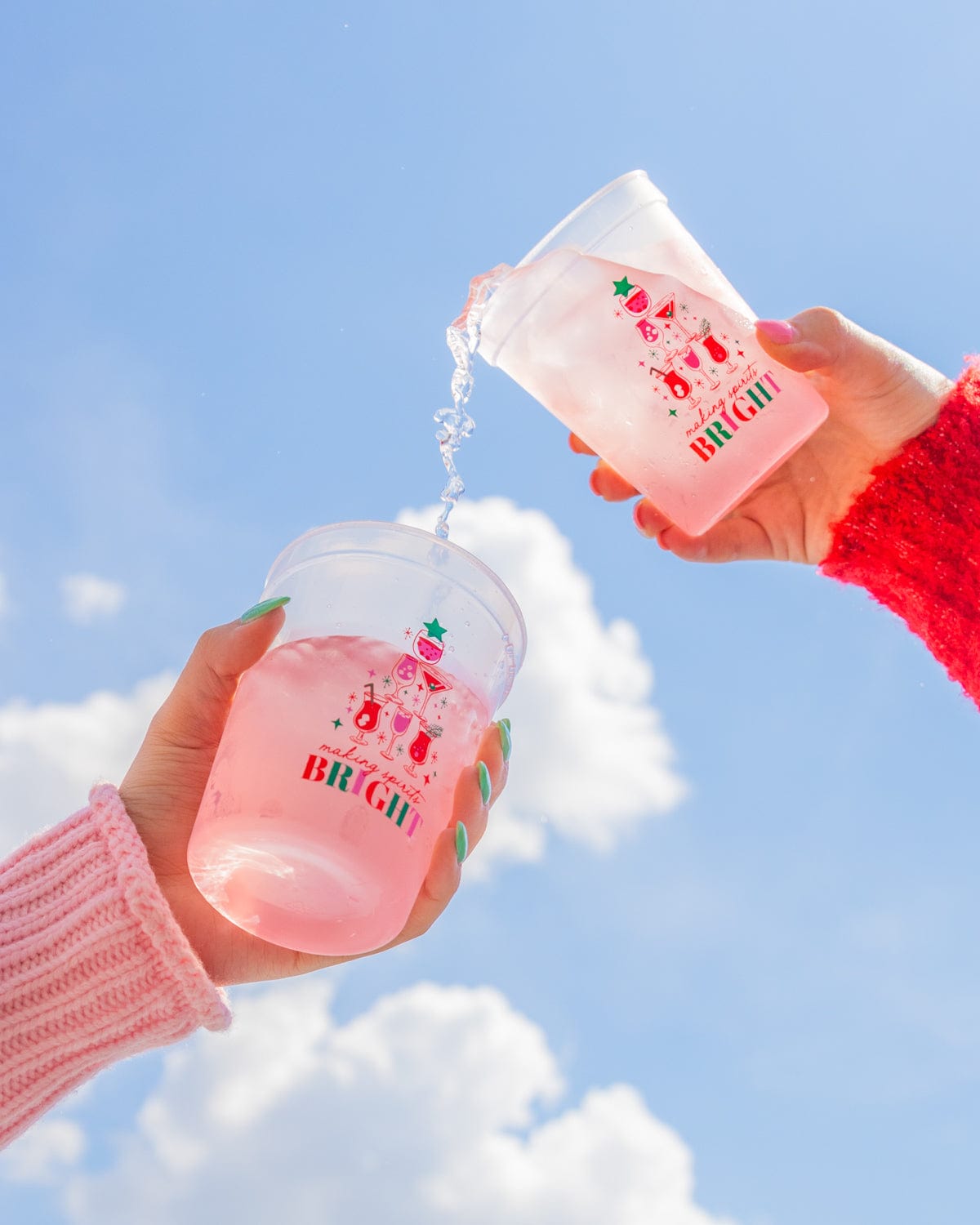 Hands holding 'Making Spirits Bright' color changing cups in sky with clouds in background. 