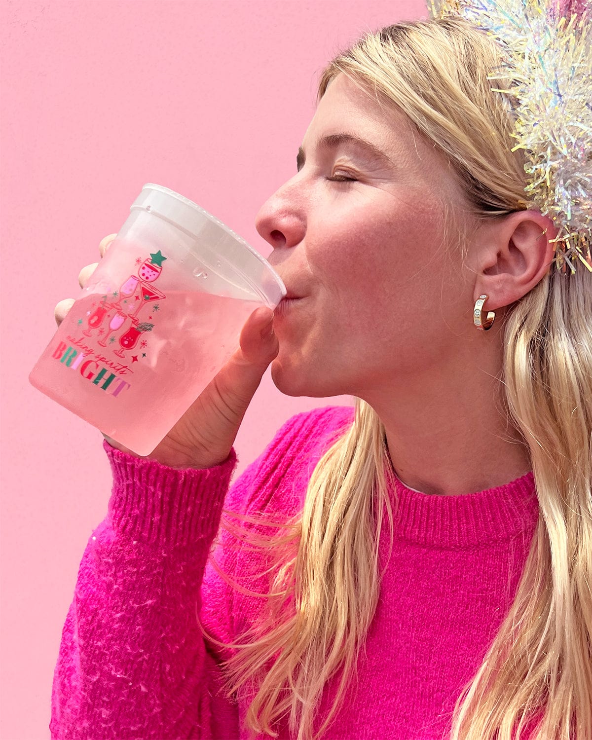 Woman sipping liquid from 'Making Spirits Bright' shot against pink background. 