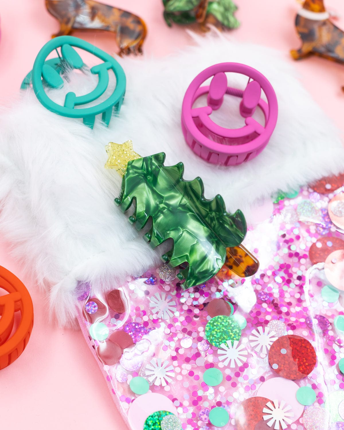 Christmas tree claw clip among assortment of holiday claw clips placed on pink confetti stocking shot against pink background. 