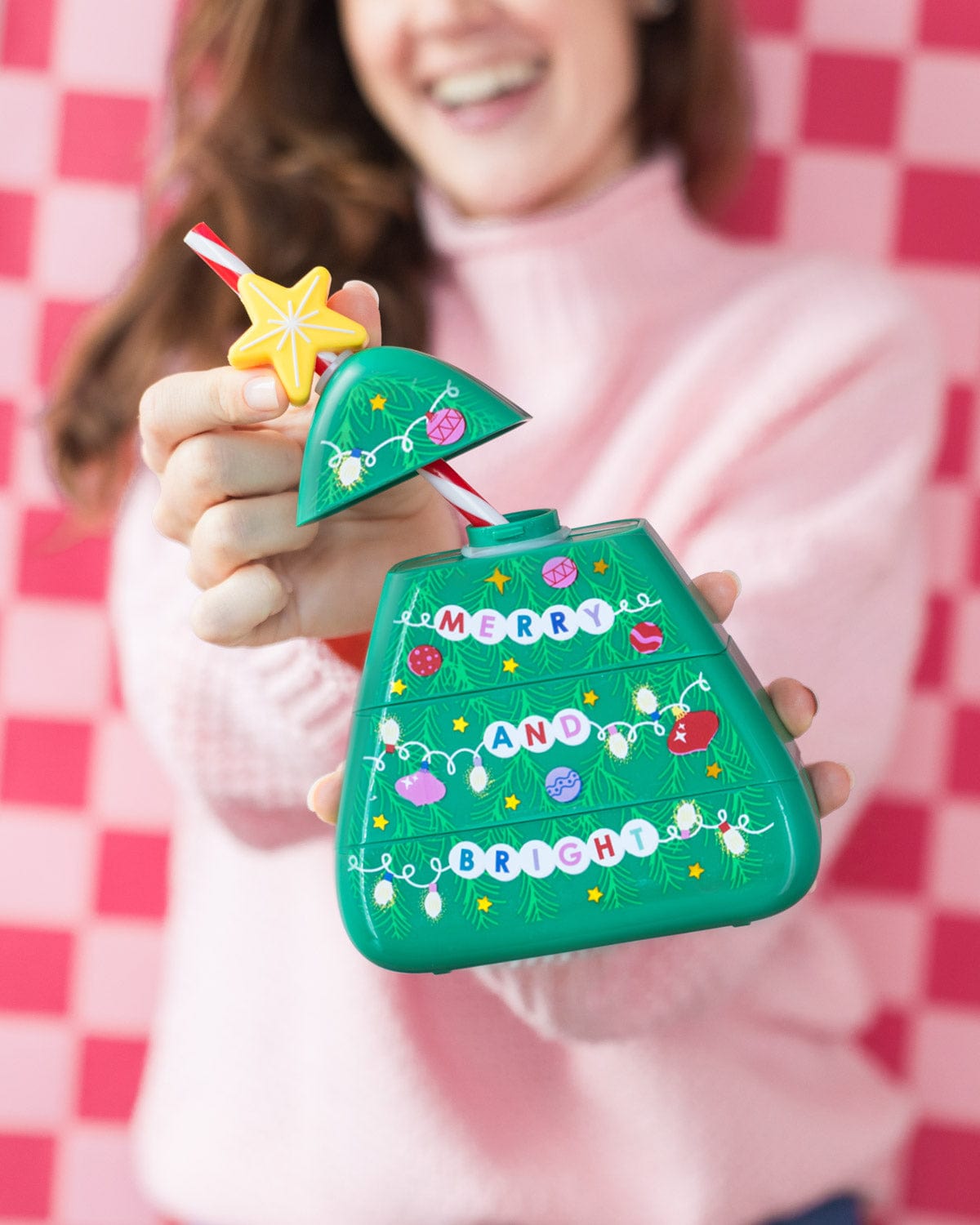 Woman holding out open Christmas Tree Sipper shot against pink checkered background. 