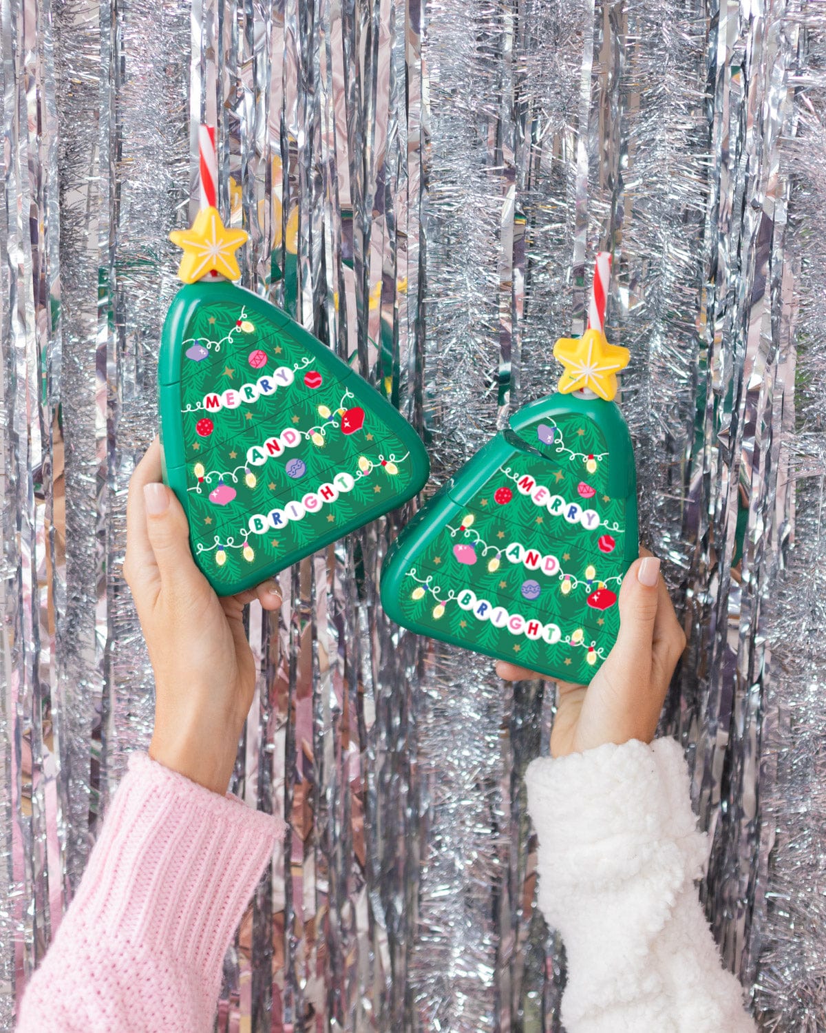 Hands holding Christmas Tree Sippers in air shot against silver tinsel background.