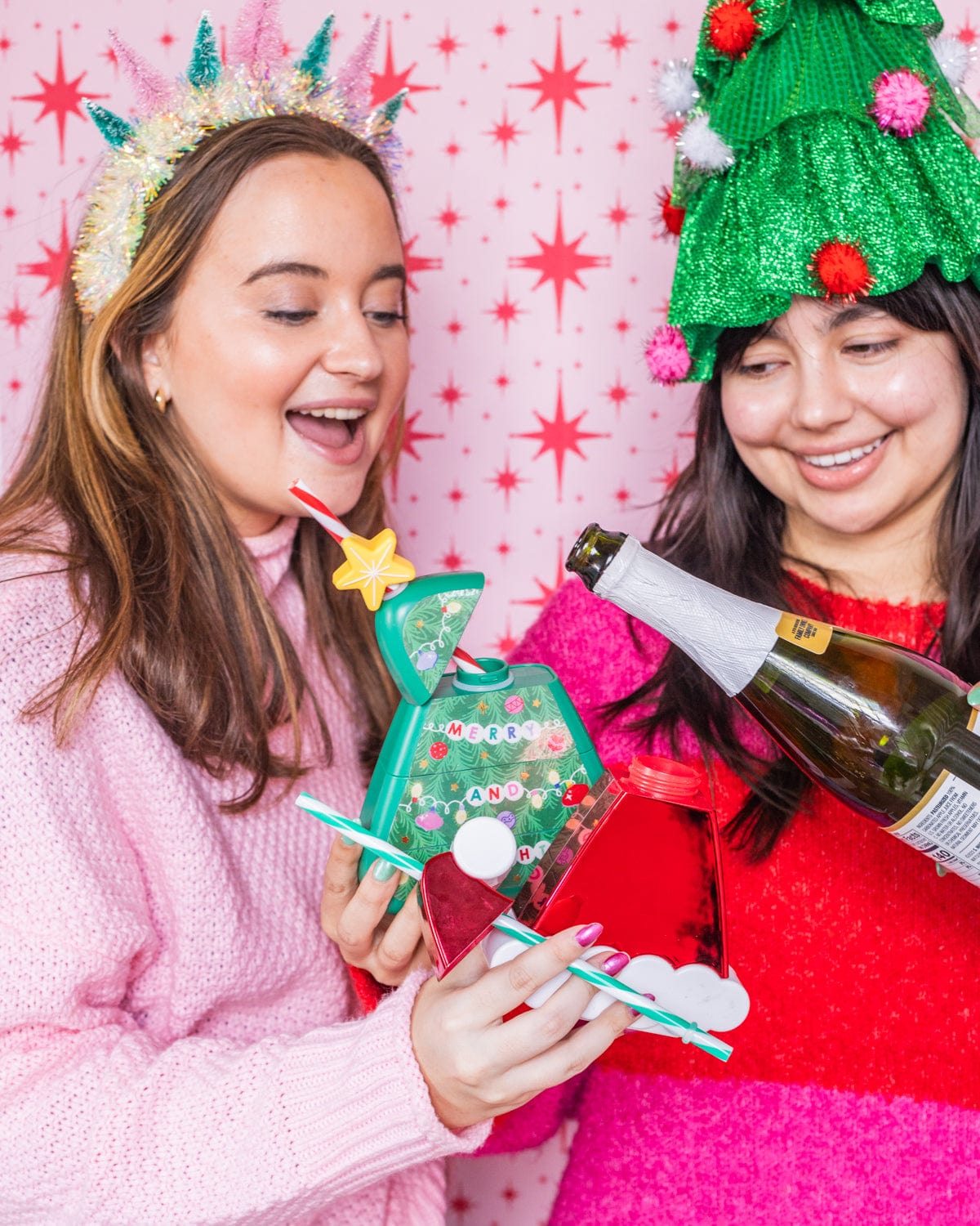 Smiling women pouring drink into Christmas Tree Sipper and Santa Hat Sipper shot against red and white patterned wallpaper. 
