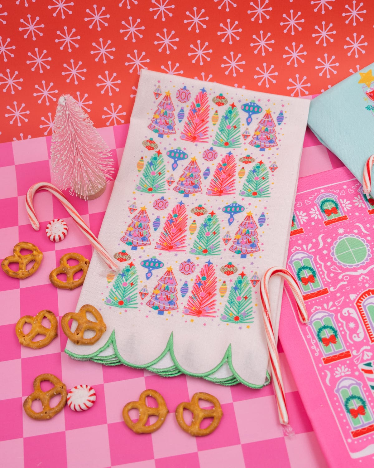 White Christmas tree tea towel surrounded by holiday treats and pink and blue holiday tea towels shot against pink checkered background. 