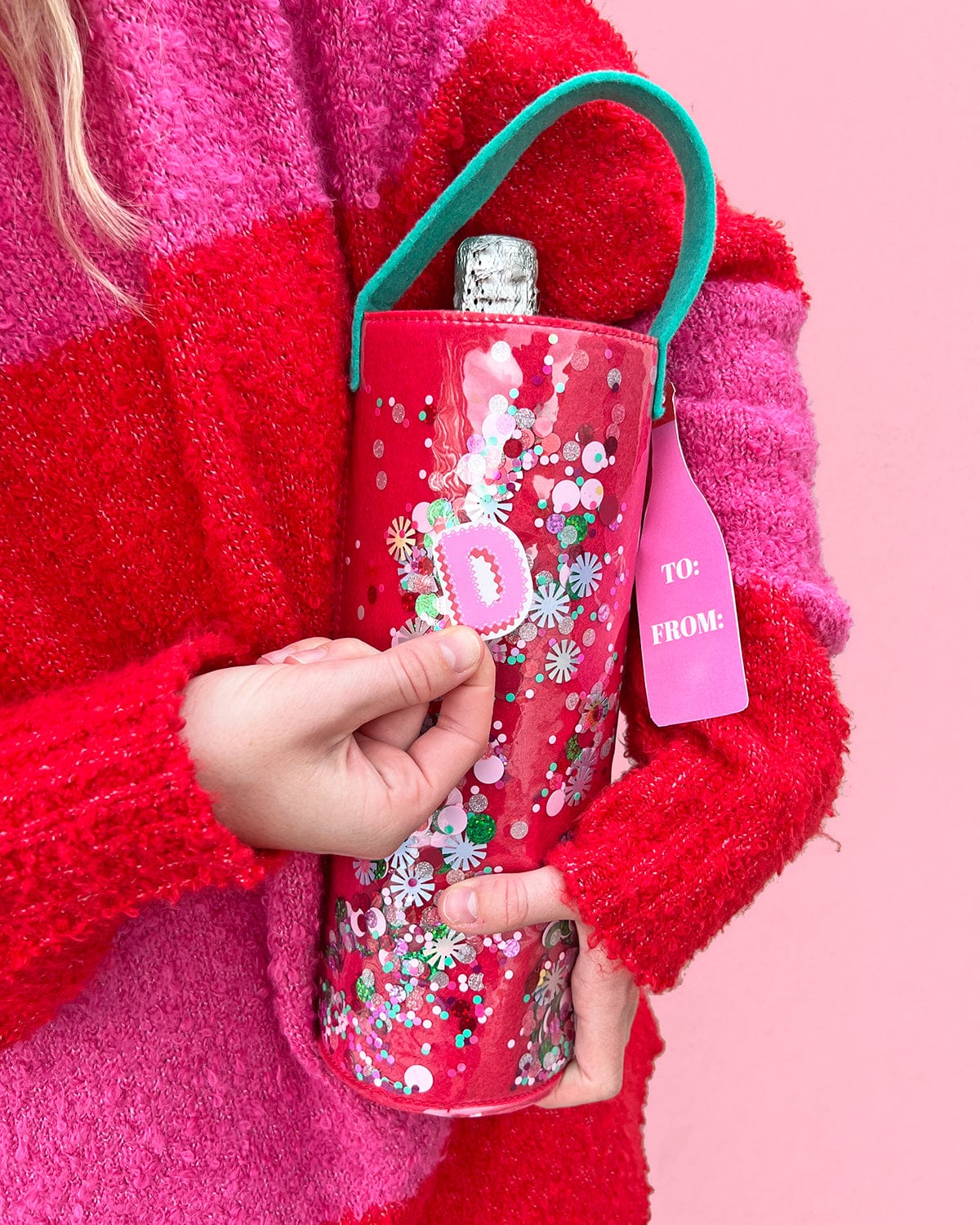 Women placing sticker onto trapped confetti wine bag shot against pink background. 