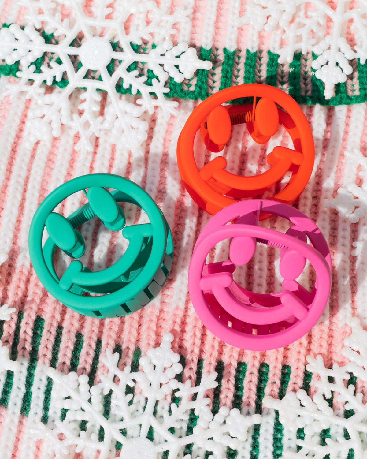 Green, red, and pink smiley claw clips surrounded by snowflakes shot against patterned fabric. 