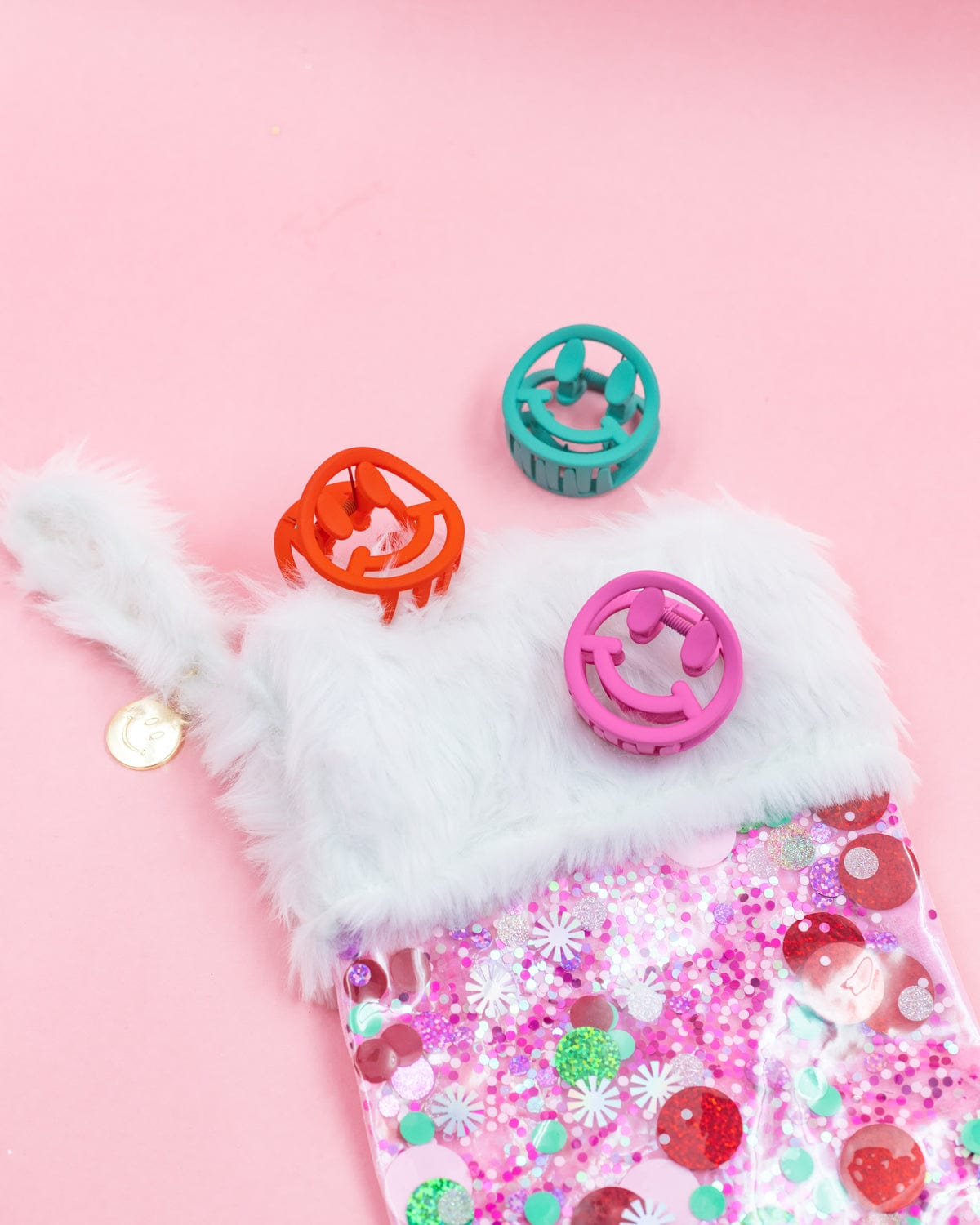 Red, green, and pink smiley claw clips placed on pink confetti stocking shot against pink background. 