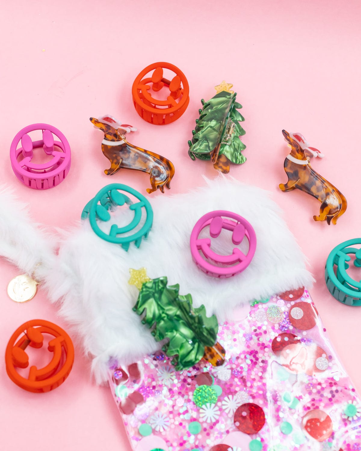 Assortment of holiday claw clips placed on pink confetti stocking shot against pink background. 