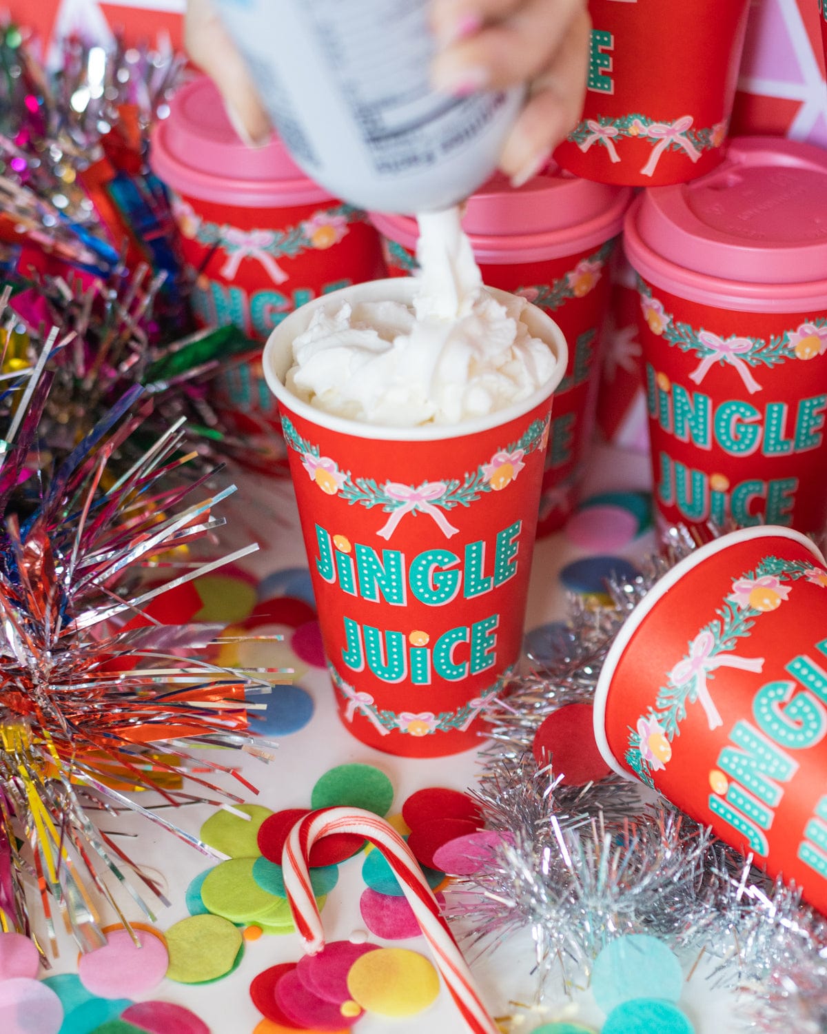Hand placing whipped cream into red 'Jingle Juice' to-go cup surrounded by assortment of confetti. 