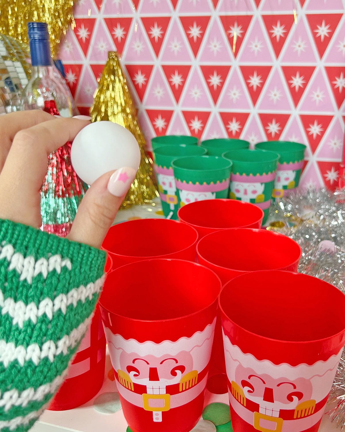 Hand holding pong style ball with green and red cups set up in background with assortment of decor. 
