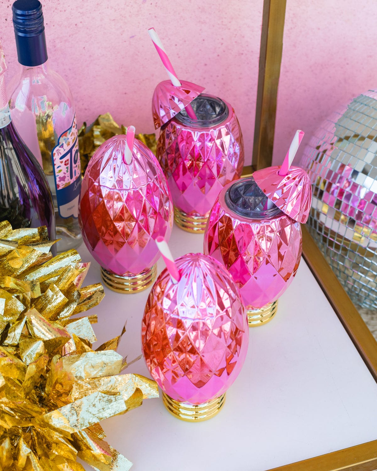Four pink faceted light bulb sippers placed on white and gold bar cart with surrounding decor. 