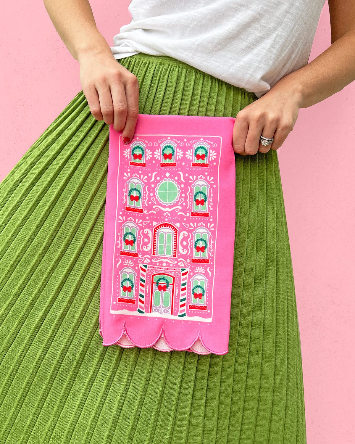 Woman holding pink gingerbread house tea towel shot against pink background. 