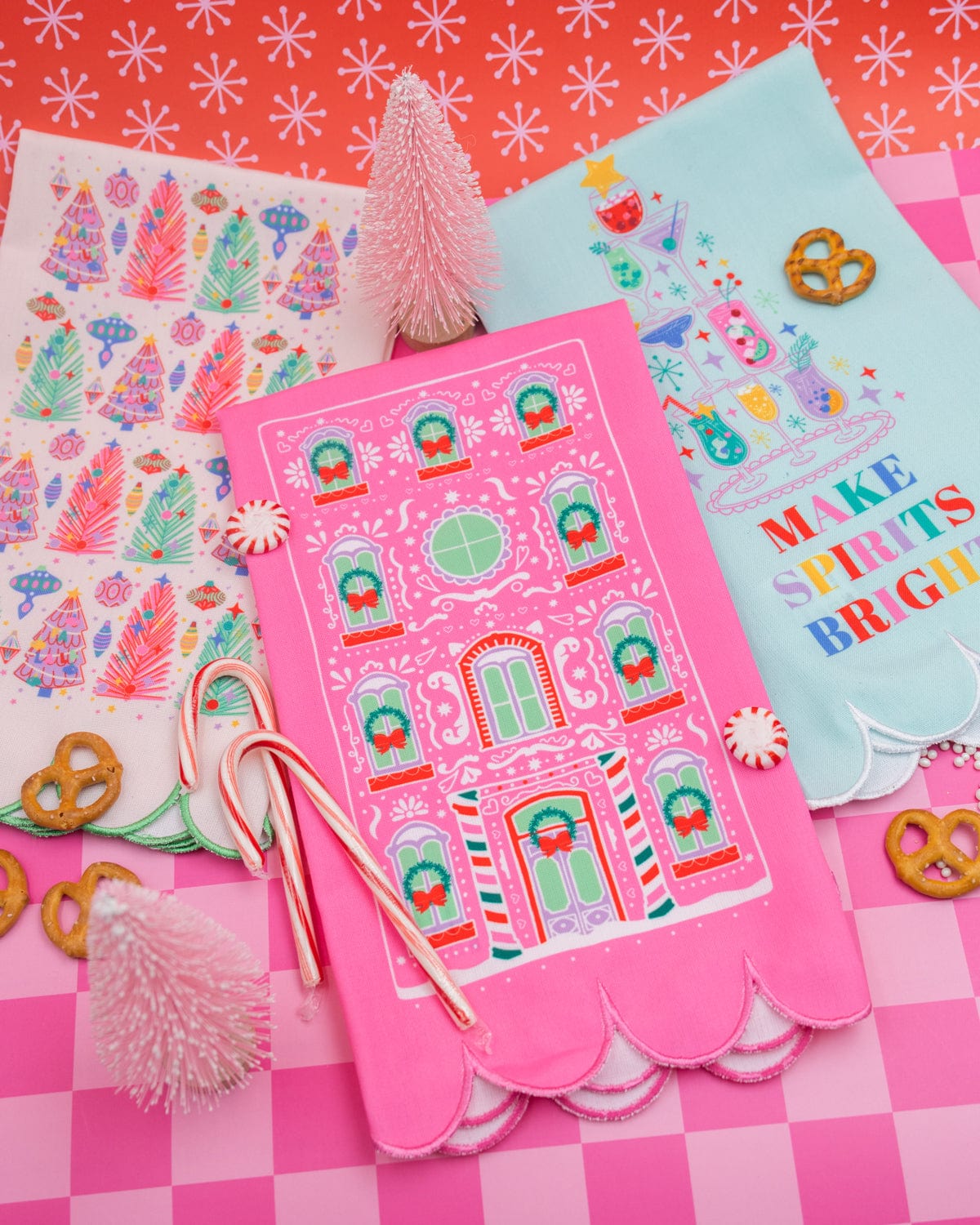 Pink gingerbread house tea towel placed with other holiday tea towels shot on pink checkered background. 