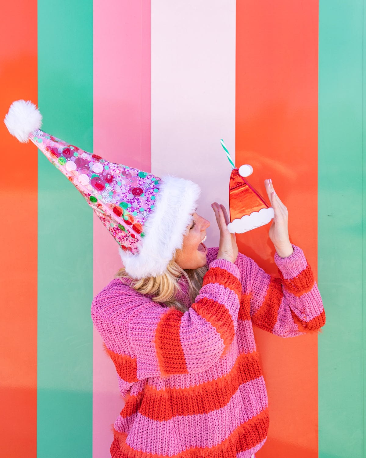 Woman holding Santa Hat Sipper while wearing pink confetti Santa Hat shot against striped colorful wall. 