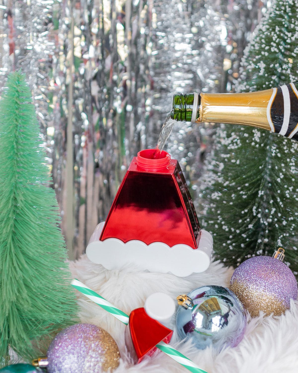 Champagne being poured into Santa Hat Sipper with holiday decor shot against silver tinsel background. 