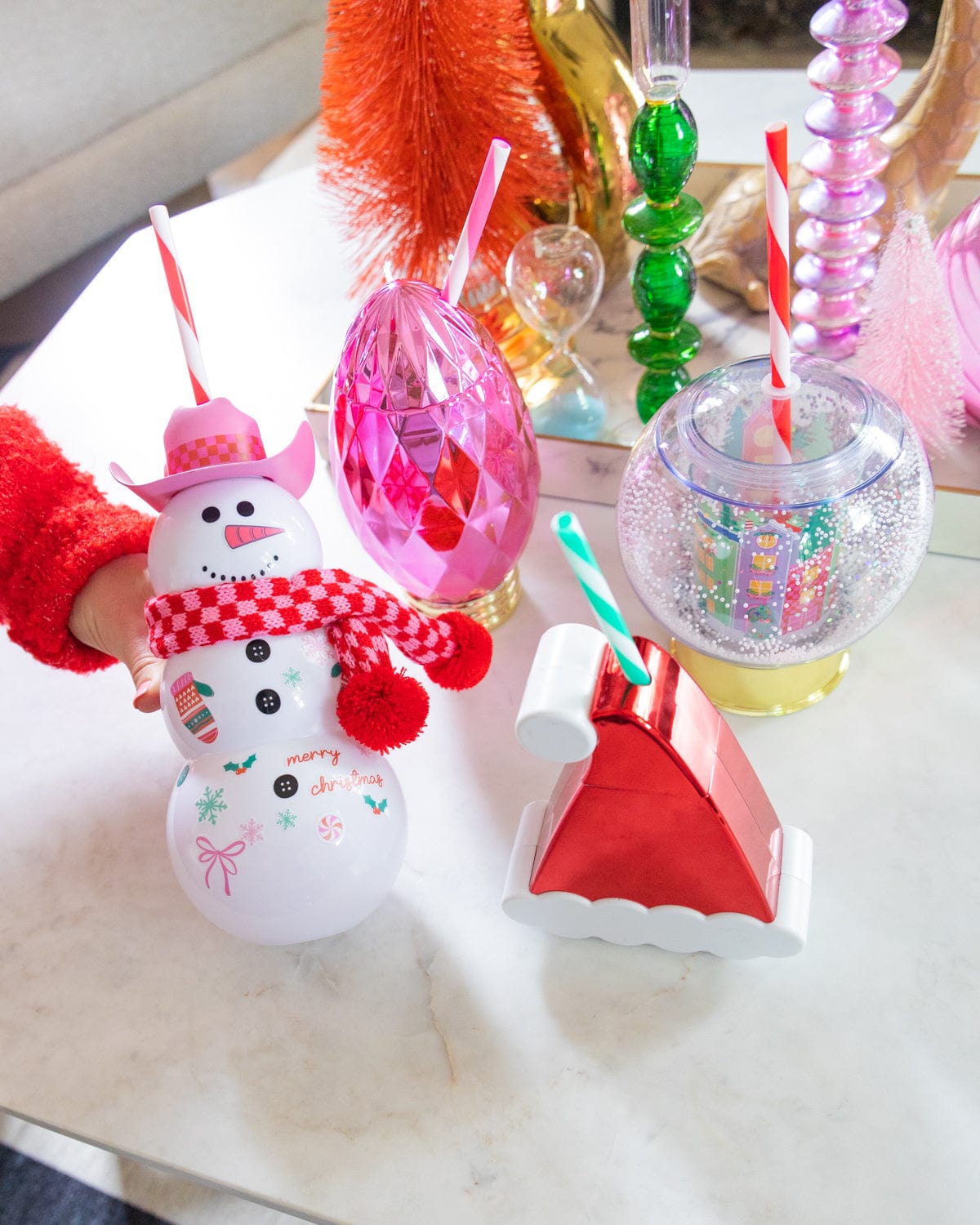 Hand holding Snowman Sipper surrounded by an assortment of holiday sippers and decor shot against a white background. 