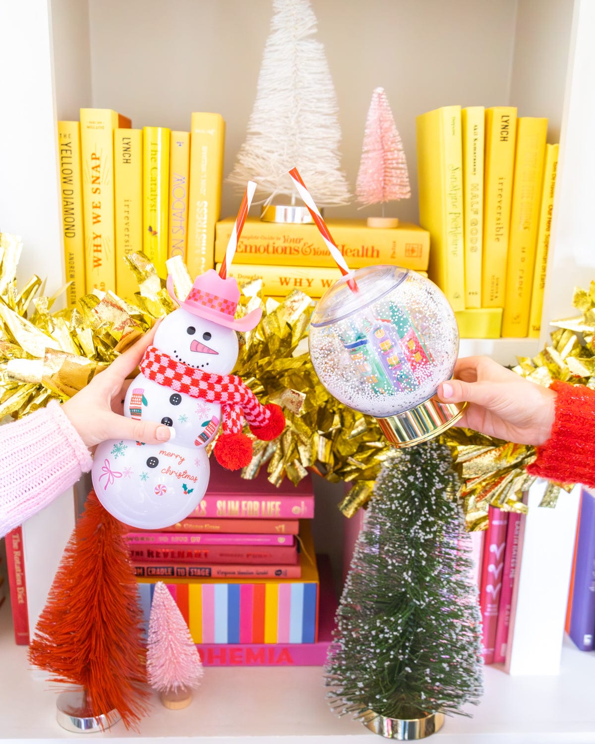 Hands holding Snowman and Snow Globe sipper in air shot against bookshelf background. 