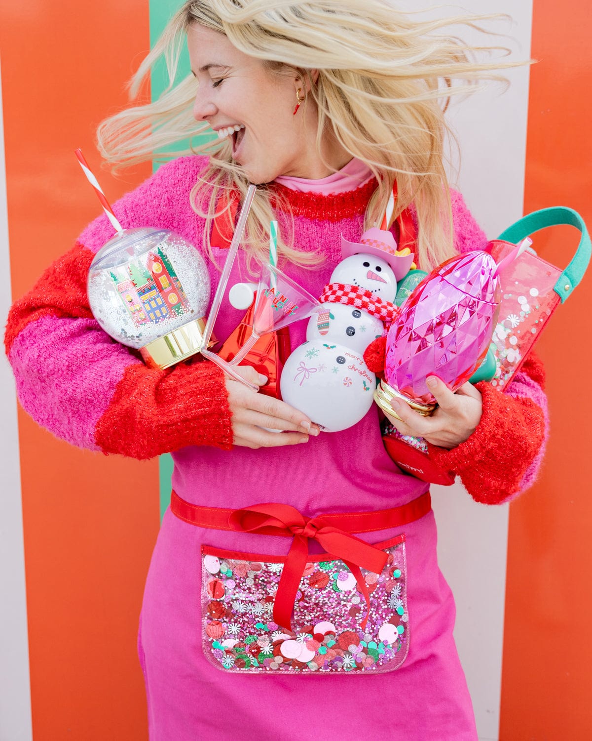 Woman holding assortment of holiday sippers and wearing pink apron shot against striped background. 