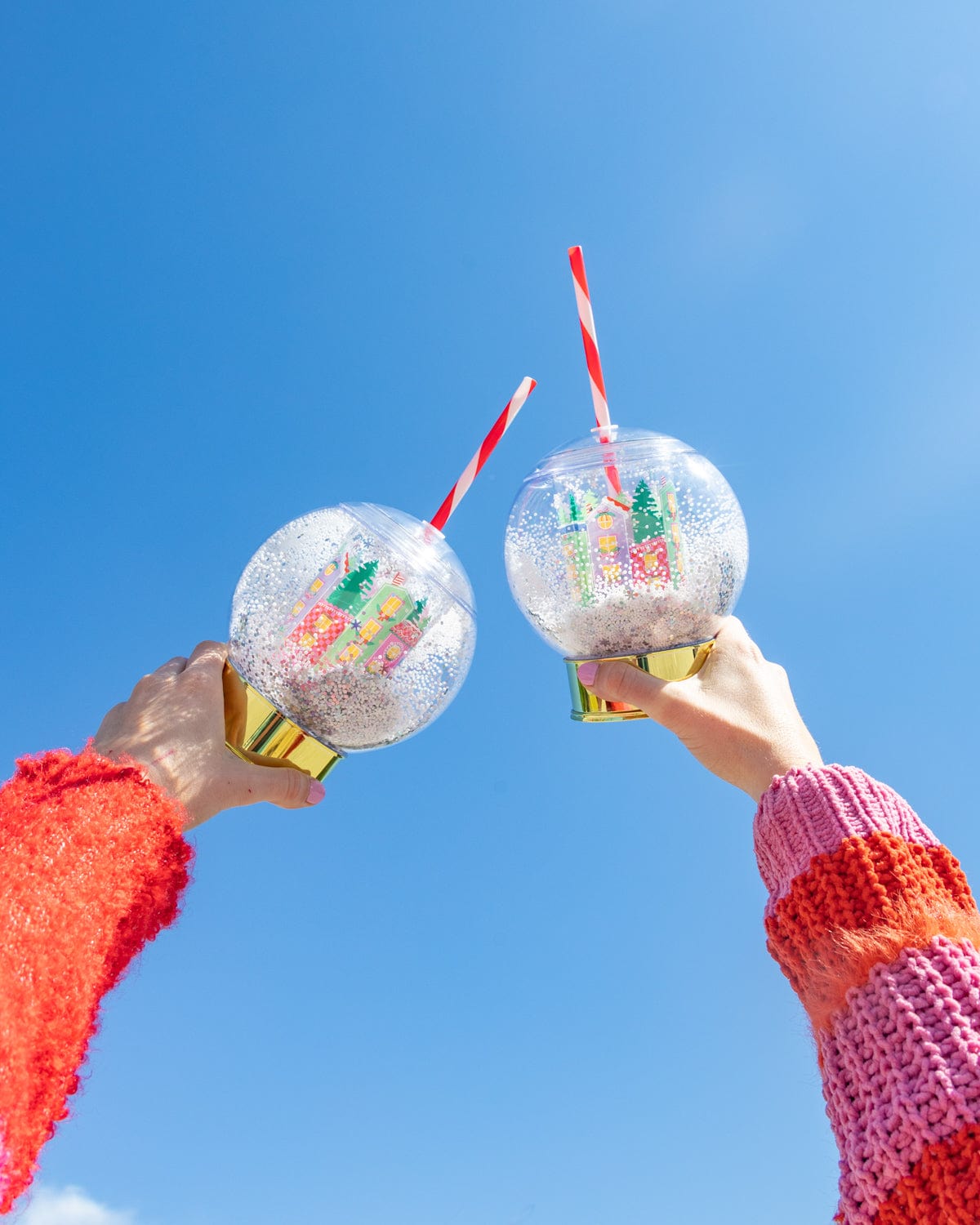 Hands holding village snow globe sippers in air shot against sky background. 