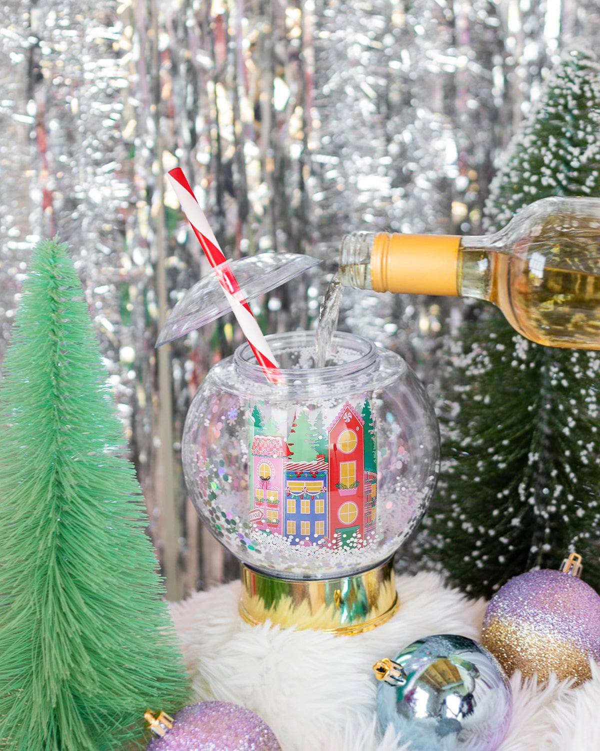 Drink being poured into village snow globe sipper surrounded by holiday decor and shot against silver tinsel background. 