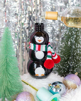 White wine pouring into black penguin sipper. Wintry background with green bottle brush trees and multicolor ornaments. 