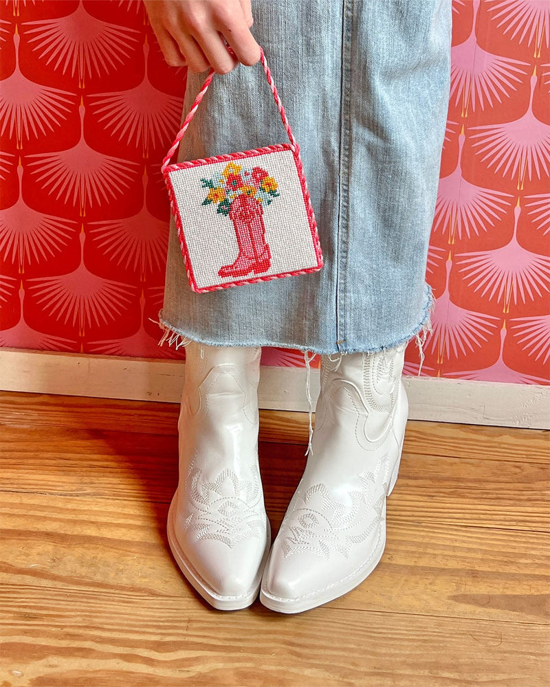 girl holding needlepoint craft in front of her denim skirt and white cowgirl boots
