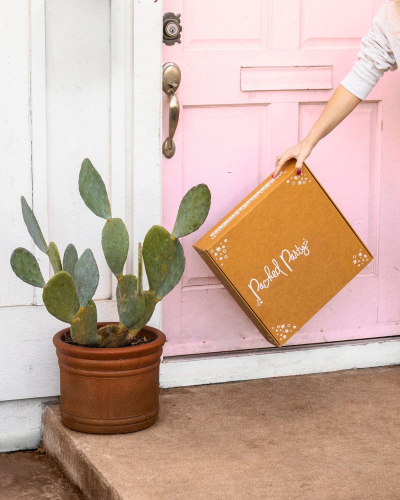 Woman picking up Packed Party Party Picks subscription box  on doorstep in front of light pink door next to cactus