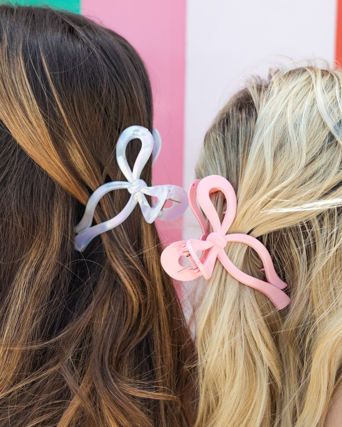 Two women stand back to back, with the photo only show the back of their heads. On the left, the person with brown hair is wearing a white bow claw clip and on the right, the blonde is wearing a pink claw clip. 