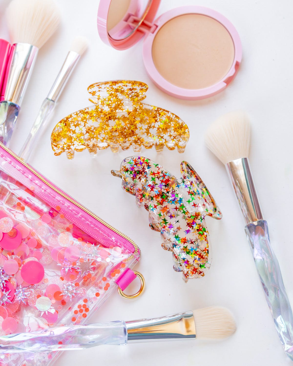 A clear vinyl pouch filled with pink confetti sits on a desk. Makeup products and accessories spill out of it, and two claw clips sit next to it. One claw clip is filled with gold stars while the other is filled with rainbow stars.